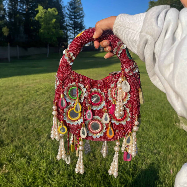 Elegant red beaded bag adorned with intricate mirror work, colorful embroidery, and pearl tassels. This traditional Indian clutch is perfect for weddings, bridal functions, and festive celebrations. With its beaded handle and vibrant design, it complements lehengas, sarees, and other ethnic outfits beautifully. A stunning accessory for brides, bridesmaids, and guests looking for a statement piece with cultural charm.