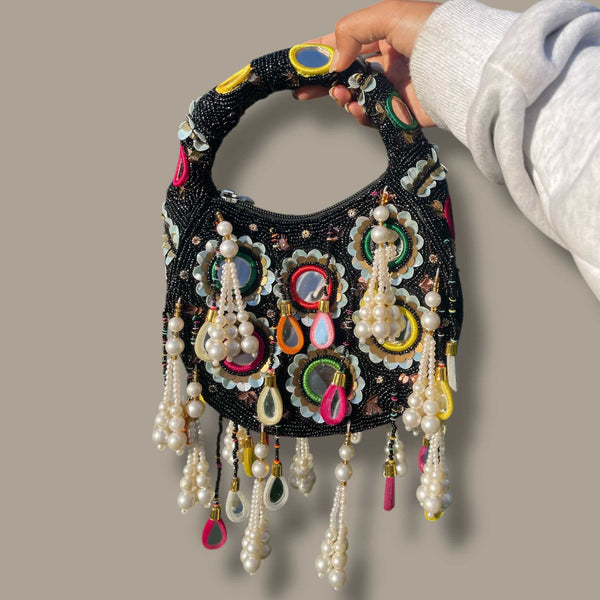 Handcrafted black potli bag featuring mirror work, colorful embroidery, and pearl tassels. This elegant Indian clutch is perfect for weddings, festivals, and traditional events. The beaded handle adds charm, making it an ideal accessory for sarees, lehengas, and bridal outfits. A must-have ethnic handbag for brides and partygoers seeking a stylish and cultural statement piece.