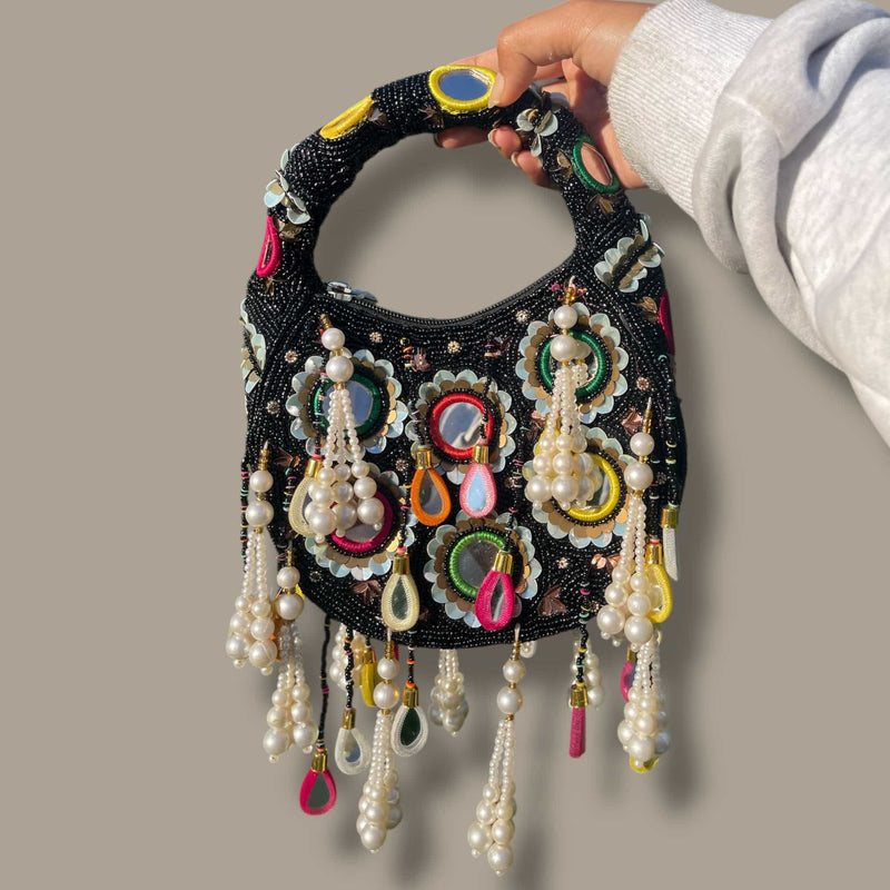 Handcrafted black potli bag featuring mirror work, colorful embroidery, and pearl tassels. This elegant Indian clutch is perfect for weddings, festivals, and traditional events. The beaded handle adds charm, making it an ideal accessory for sarees, lehengas, and bridal outfits. A must-have ethnic handbag for brides and partygoers seeking a stylish and cultural statement piece.
