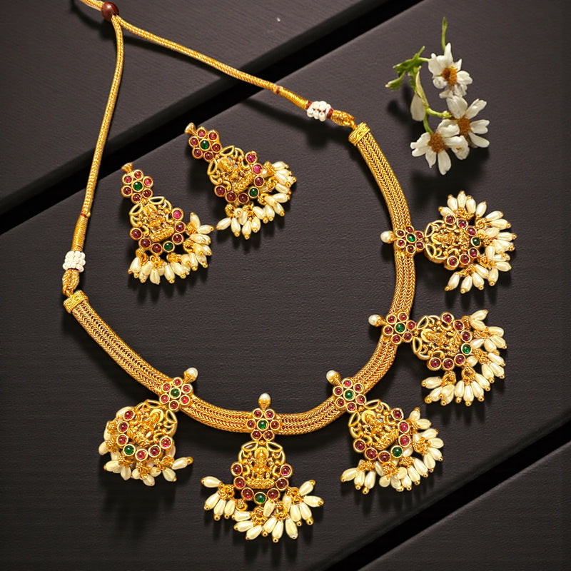 The image shows a beautifully arranged South Indian matte antique gold temple jewelry set displayed on a dark charcoal-black textured surface. The composition features a traditional Lakshmi temple necklace paired with matching earrings, styled in an elegant flat-lay format. The deep black background enhances the warm glow of the matte antique gold finish, making the jewelry appear rich, vibrant, and luxurious.
The necklace is crafted in a woven gold design, with a thick braided chain that curves gracefully 