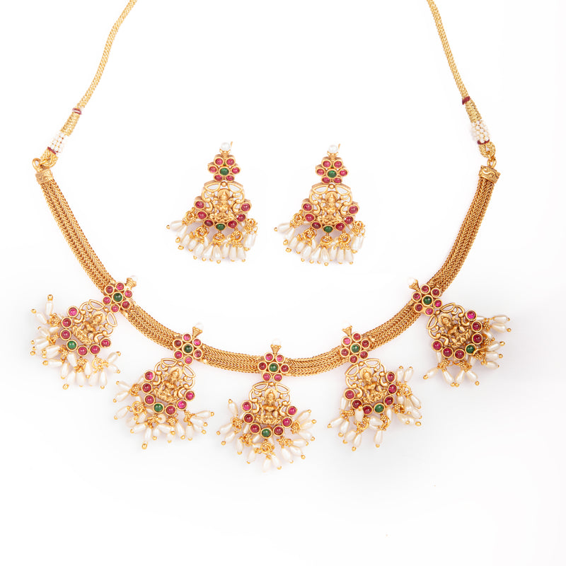 The image displays a complete South Indian matte antique gold Lakshmi temple jewelry set arranged neatly on a bright white background. The set includes a traditional temple-style necklace and a matching pair of earrings, both designed with intricate craftsmanship that highlights the cultural heritage of South Indian jewelry. The necklace is placed in a gently curved shape, showing its full structure, while the earrings are positioned above it, centered and symmetrical, creating a balanced and visually appea