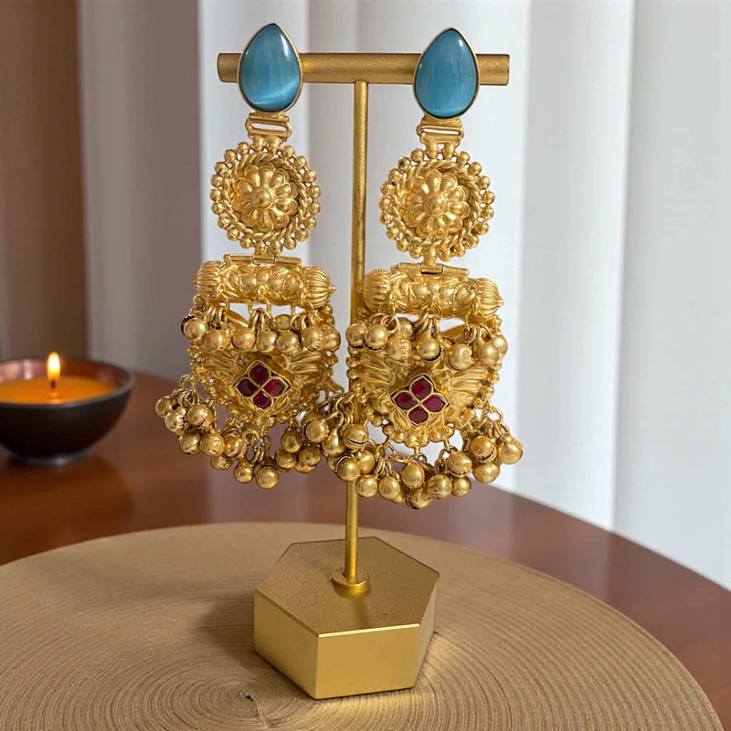 Gold earrings with blue stones on a gold stand on a wooden surface. 92.5 silver gold-plated temple earrings displayed on a jewelry stand, captured from a close-up angle. The earrings feature ruby stones, floral detailing, and hanging ghungroos that highlight their traditional South Indian craftsmanship. The antique gold finish and intricate handwork are visible from this perspective, making them perfect for bridal, festive, and ethnic wear photography or product display.