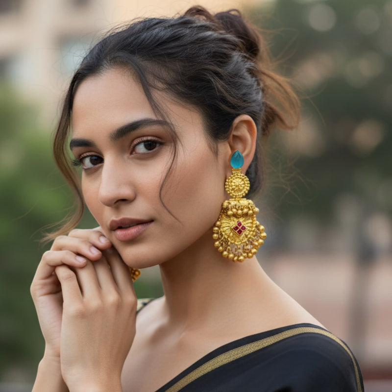 Woman wearing large, ornate gold earrings with a blurred background. Model wearing antique gold 92.5 silver temple earrings featuring intricate floral motifs, ruby stones, and traditional ghungroos (bells). The earrings have a rich antique gold plating and detailed craftsmanship, adding elegance and cultural charm. Perfect for South Indian bridal looks, festive celebrations, or traditional events. These handcrafted earrings beautifully complement sarees, lehengas, and ethnic outfits for an elegant statement