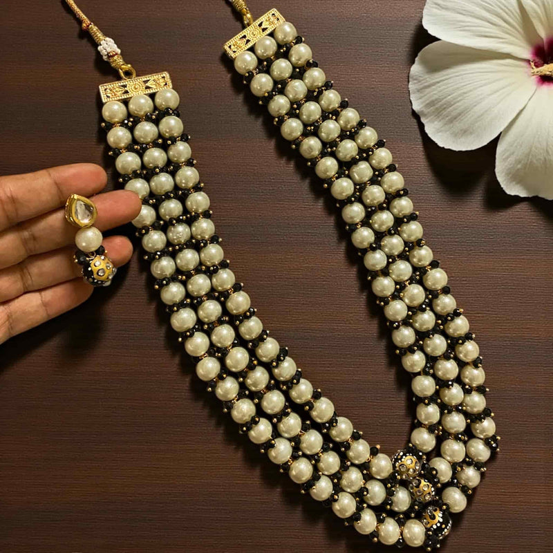 Close-up image of the Fatiha Long Pearl Necklace with Earrings, a handcrafted multi-layer Indian pearl mala made with ivory pearls, black beads, Kundan stones, and Meenakari gold-plated detailing. Displayed on a dark wooden surface beside a white flower, with the matching Kundan pearl earring shown in hand. Ideal for bridal wear, festive outfits, and traditional Indian jewelry styling.