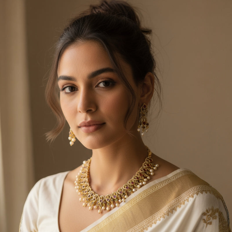 Model showcasing a traditional gold necklace adorned with CZ, ruby, and emerald stones in an intricate design. The necklace sits beautifully around her neck, accentuated by delicate pearl drops that add a touch of grace. The gold finish glows against her skin, enhancing the elegance of her bridal or festive attire, perfectly capturing the essence of South Indian jewelry craftsmanship.