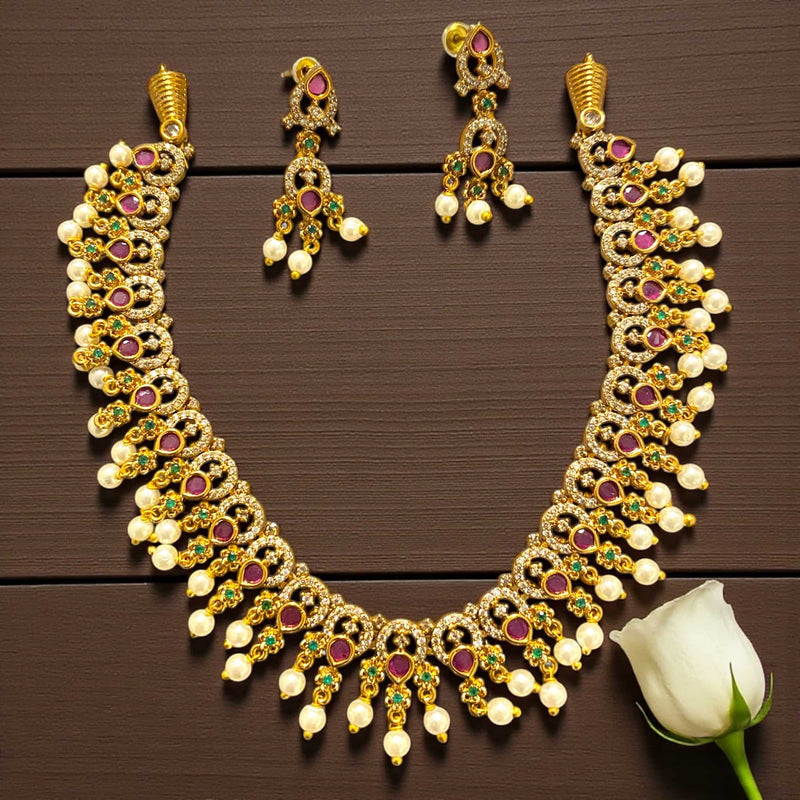 Gold necklace set with matching earrings featuring intricate CZ, ruby, and emerald stone design in a rich gold finish. The necklace is adorned with pearl drops and detailed craftsmanship, giving it a luxurious South Indian traditional look. Perfect for brides and festive occasions, this ethnic jewelry set exudes elegance and timeless charm, displayed on a wooden background beside a white rose.