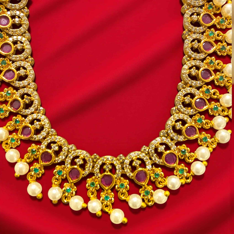 Gold necklace featuring intricate craftsmanship with CZ, ruby, and emerald stones set in a rich gold-plated base. The design includes repeating motifs adorned with sparkling stones and elegant pearl drops. Displayed on a wooden background with a white rose, this traditional South Indian necklace exudes timeless beauty, perfect for weddings, bridal wear, and festive celebrations.