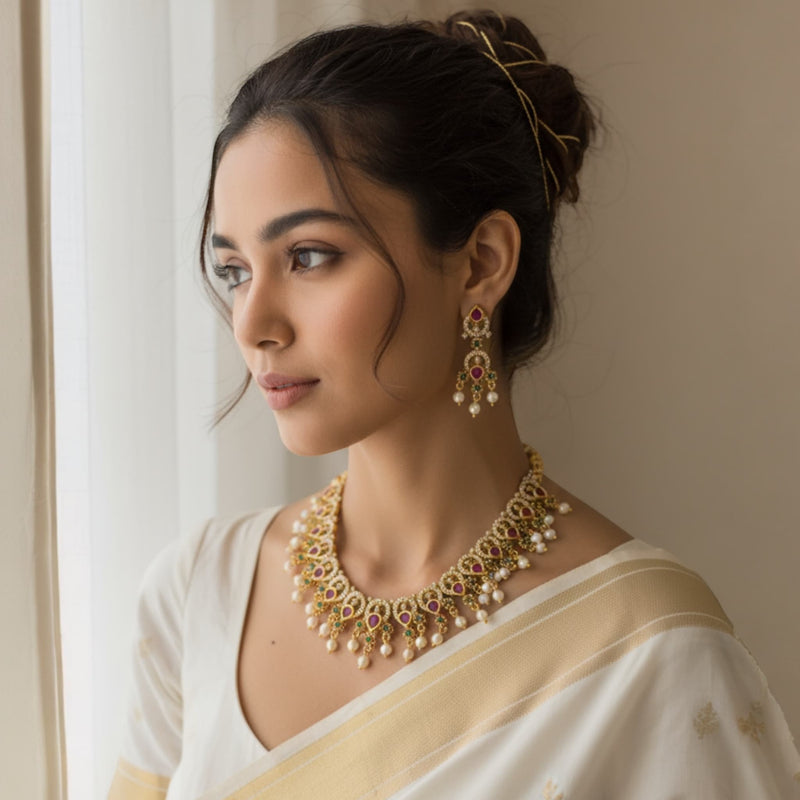 Woman wearing traditional jewelry including a necklace and earrings, standing against a neutral background. Model wearing a gold necklace featuring intricate CZ, ruby, and emerald stones in a traditional South Indian design. The necklace rests gracefully on her neckline, highlighting its pearl drops and fine gold detailing. The rich colors of the stones complement her outfit and makeup, creating an elegant and regal bridal look perfect for weddings and festive occasions.