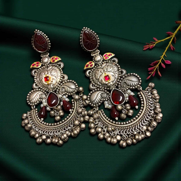 German silver chandbali earrings with antique oxidized finish, deep ruby stones, carved motifs, peacock-inspired detailing, small pearl accents, and dangling ghungroo beads. Traditional Indian ethnic jewelry with intricate craftsmanship, bold statement design, and rich detailing. Perfect for weddings, festive wear, cultural events, and special occasions. Detailed close-up of the earrings.
