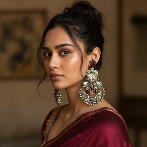Woman wearing German silver chandbali earrings with ruby stones, antique oxidized finish, floral motifs, peacock design, and ghungroo detailing. Traditional ethnic Indian jewelry styled for festive and wedding looks.