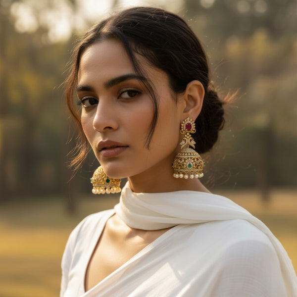 Woman wearing gold CZ jhumka earrings featuring ruby stone studs, emerald accents, and delicate pearl hangings. The earrings have intricate gold filigree and sparkling cubic zirconia, styled beautifully with traditional attire. Perfect accessory for weddings, festive events, and elegant South Asian fashion.