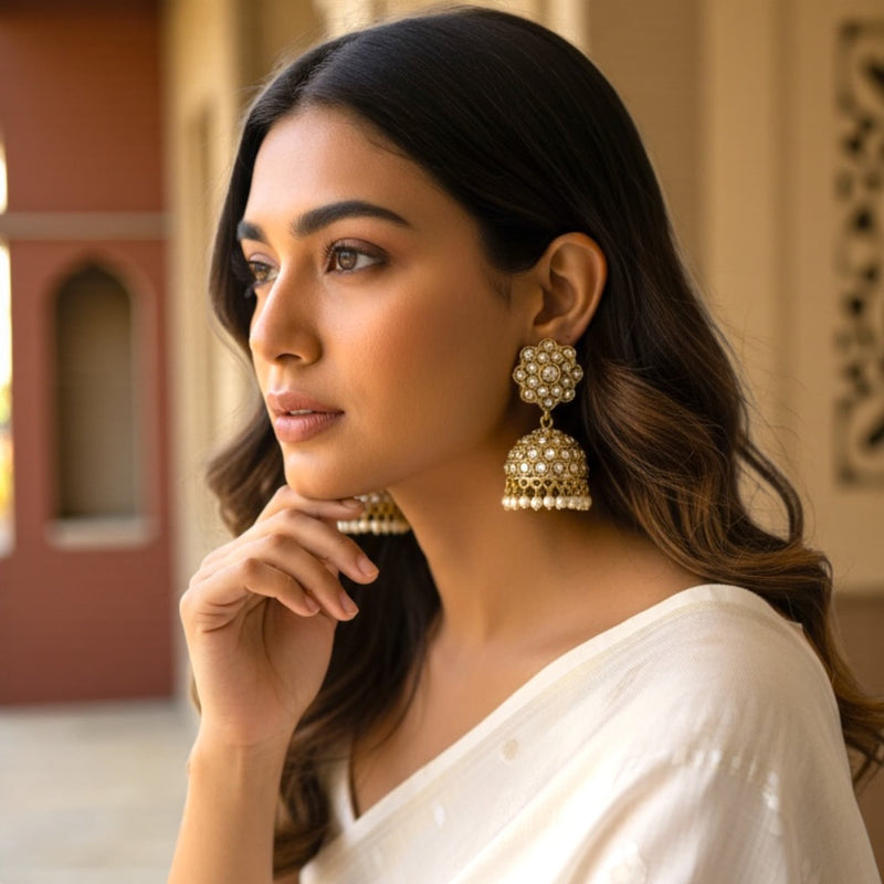 A woman wearing antique mehndi gold Polki jhumka earrings with pearl drops, shown in soft natural light. The earrings feature a floral Polki stud and an ornate dome-shaped jhumka with intricate detailing. She is dressed in an elegant off-white saree, and the warm, blurred architectural background highlights the traditional design, shine, and craftsmanship of the jhumkas.