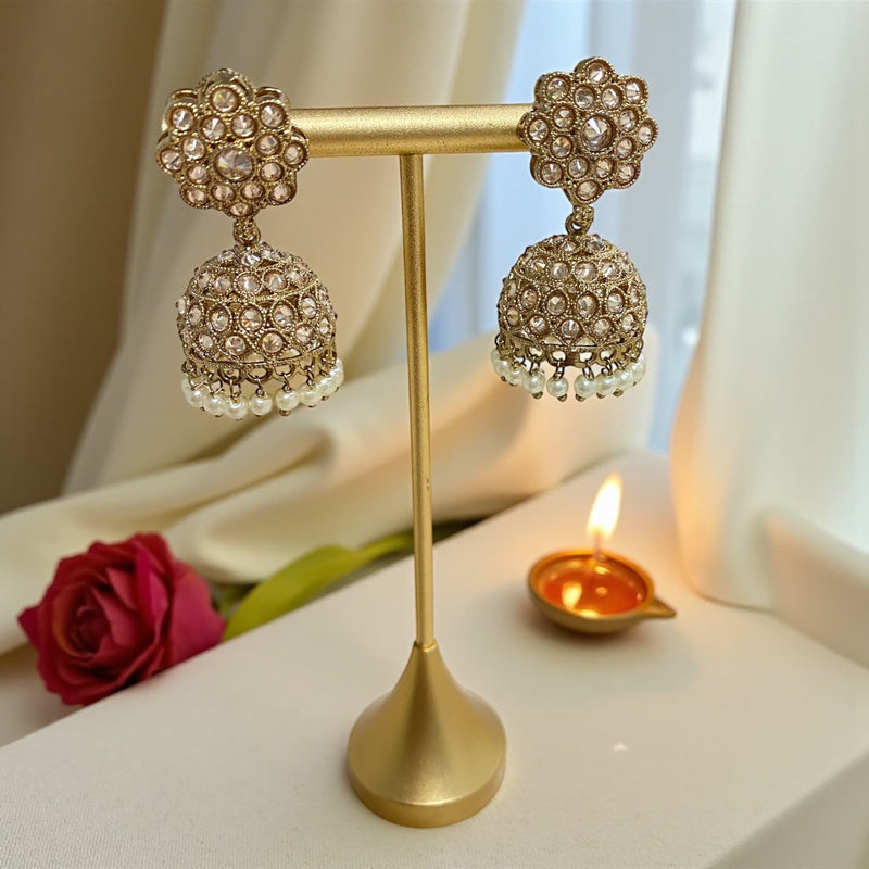 Antique mehndi gold Polki jhumka earrings with pearl drops displayed on a gold stand. The floral Polki studs and detailed dome-shaped jhumkas are shown in soft lighting, with a red rose, cream fabric, and a lit diya in the background, creating a festive and elegant setting that highlights the craftsmanship and sparkle of the jewelry.